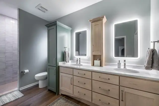 Ensuite bathroom remodel in DeForest, WI with dual wood vanities, backlit mirrors and a green linen tower